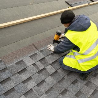 Qualified workman in uniform work wear using air or pneumatic nail gun and installing asphalt or bitumen shingle on top of the new roof under construction residential building